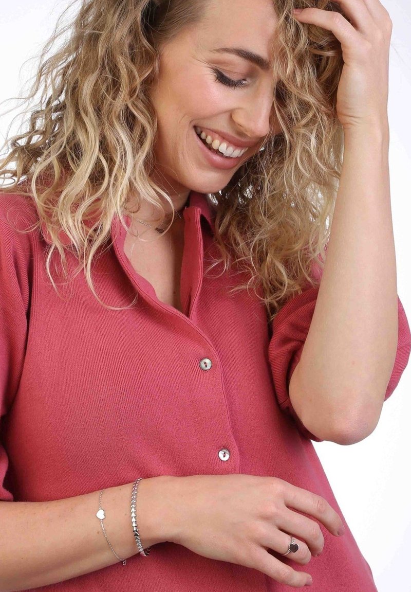 Button-up shirt in pink fabric, featuring a collar and short sleeves. Silver bracelets adorn a wrist, with a minimalist design and heart charm.