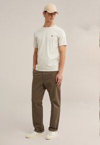 Krémové tričko s malým logem, hnědé chinos kalhoty a bílé tenisky. Model má na hlavě béžovou kšiltovku a stojí proti neutrálnímu pozadí.