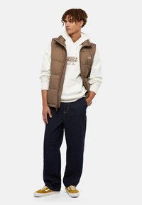 Bruine gewatteerde vest over een witte hoodie met een logo, gecombineerd met donkerblauwe wijde jeans en gele sneakers met witte veters.
