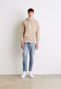 Sudadera beige con capucha y logo, combinada con jeans de mezclilla azul claro y zapatillas blancas. Tejido suave, con un corte relajado y bolsillo frontal.