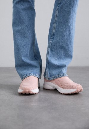 Personne portant un jean bleu clair et des baskets roses en mesh sans lacets, debout sur un sol gris.