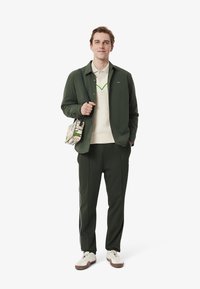 Grön knäppjacka över en krämfärgad väst, matchad med gröna byxor och vita sneakers. En mönstrad väska hänger över axeln.