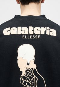 Πίσω μέρος μαύρου πουκαμίσου με το κείμενο «gelateria ELLESSE» πάνω από ένα γραφικό με ένα χέρι που κρατάει μια χωνάλα με παγωτό.