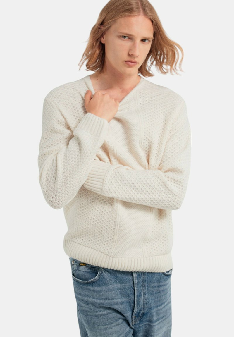 Witte gebreide trui met een textuurpatroon en ribbels aan de mouwen, gedragen over lichtblauwe spijkerbroek. Het ontwerp heeft een V-hals en een ontspannen pasvorm.