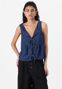Jeanstopp uten ermer i dyp blå, med V-hals, snor detalj i midjen, og en utskjært design. Kombinert med svarte bukser med knapper.