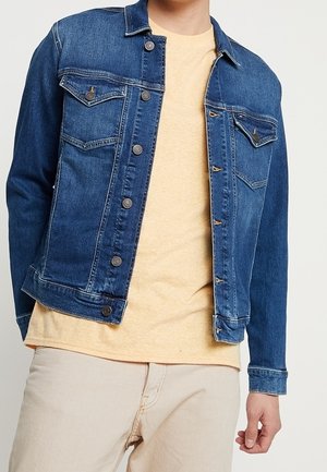 Homme portant une veste en denim bleue sur un t-shirt jaune pâle et un pantalon beige clair, debout devant un fond clair uni.