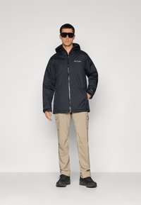 Black waterproof jacket with a hood, front zipper, and logo on the chest. Paired with beige cargo pants and black shoes.