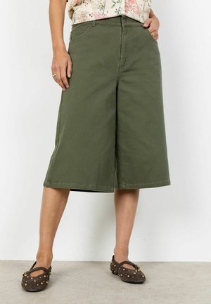 Personne portant un short large longueur genou vert olive, une chemise fleurie et des chaussures marron foncé à enfiler cloutées, debout contre un mur blanc.