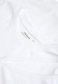 Baltos medvilninės marškinėliai su apvaliu kaklu ir lygiu audiniu. Etiketėse nurodyta prekės ženklas ir dydis. Du marškinėliai šiek tiek vienas ant kito uždėti.