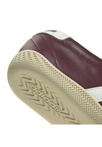 Gros plan sur le talon d'une basket en cuir marron foncé avec une semelle texturée beige montrant le logo Adidas sur la semelle et un détail de bande blanche.