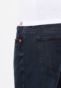 Dunkelblaue Jeans mit einem roten Akzent an der Tasche, kombiniert mit einem weißen Baumwolloberteil, das mit einem kleinen Logoetikett am Saum versehen ist.