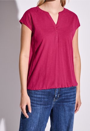 Kobieta ubrana w ciemnoróżowy krótki top zapinany na guziki i niebieskie dżinsy, stoi z lewą ręką luźno opuszczoną wzdłuż ciała.