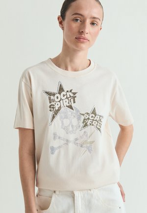 Femeie care poartă un tricou bej deschis cu un grafic de craniu și oase încrucișate și forme de stele cu cuvintele "Rock Spirit" și "Cal Pers."