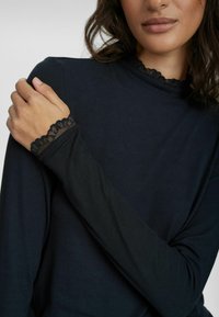 Svart ribbad långärmad topp med hög krage och spetsdetaljer vid ärmsluten. Tyget ser mjukt och figurnära ut, vilket framhäver designens detaljer.