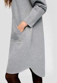 Šedé šaty s dlouhými rukávy a kulatým lemem, které mají přední kapsu. Tkanina je hladká a zdá se, že je jemná na dotek.