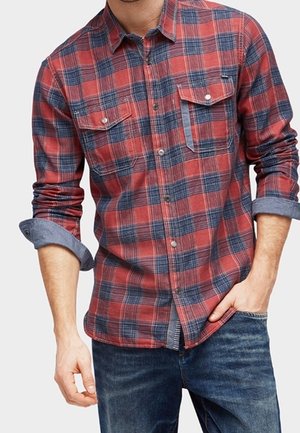 Homme portant une chemise à carreaux rouge et bleue à boutons avec manches retroussées et un jean bleu, main partiellement dans la poche.