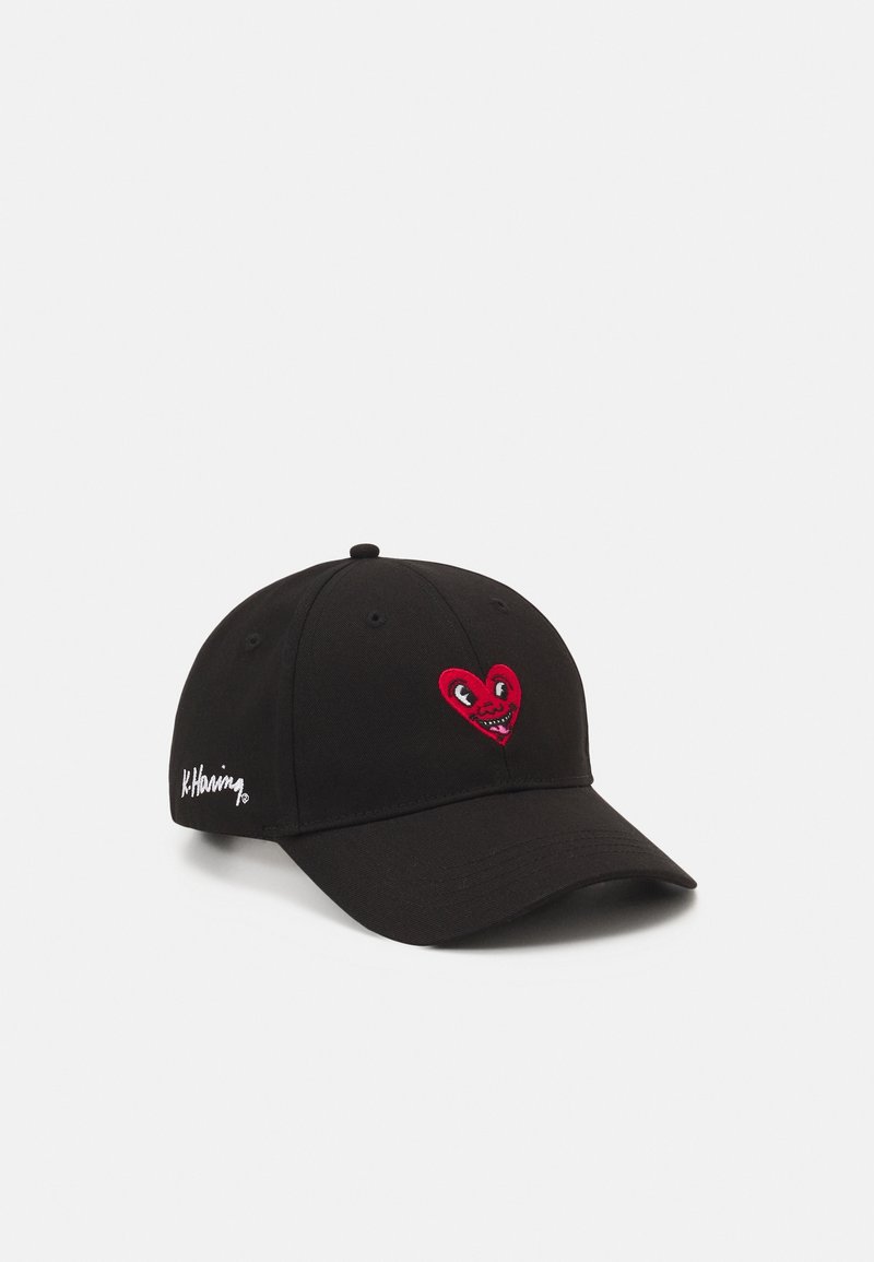 Casquette de baseball noire avec un cœur rouge présentant des yeux et une bouche à l'avant, et la signature blanche "K.Haring" brodée sur le côté.