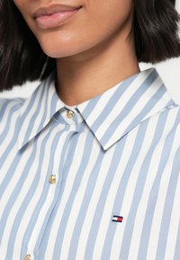 Κοντινό πλάνο μιας γυναίκας που φοράει ένα μπλε-λευκό κάθετα ριγέ πουκάμισο με μικρό ορθογώνιο λογότυπο στο στήθος.