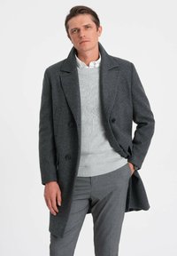 Mørk grå uldfrakke med en dobbeltbrystet design, store faldrevers, to knapper og sidelommer. Båret over en lysegrå sweater og grå bukser.