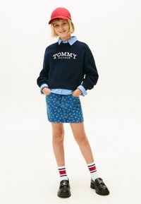 Maglione navy con il logo "TOMMY HILFIGER", camicia blu a righe sotto, gonna di jeans con stampa floreale, berretto rosso, mocassini neri e calzini a righe.