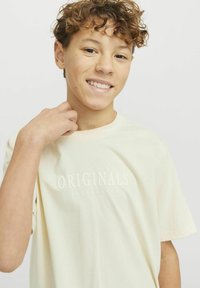 Kremasta bombažna majica s kratkimi rokavi, ki na sprednji strani nosi reliefni napis "ORIGINALS JACK & JONES". Mehka tekstura, sproščen kroj.