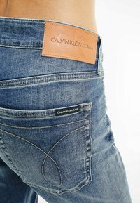 Džínové kalhoty v středně modrém odstínu s koženou etiketou Calvin Klein Jeans, kontrastním prošitím a charakteristickým zakřiveným designem kapes.