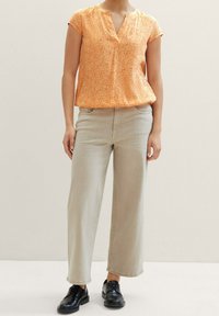 Blouse orange à manches courtes avec un motif de feuilles, associée à un pantalon large beige clair et des chaussures noires à lacets.