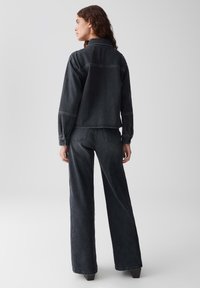 Frau in schwarzer Denimjacke und passenden hochgeschnittenen weit geschnittenen Jeans, steht mit dem Rücken zur Kamera vor einem einfarbigen Hintergrund.