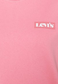 Camiseta de algodón rosa con cuello redondo y un logo blanco de Levi's sobre un fondo rojo, centrado en el pecho. Textura suave.