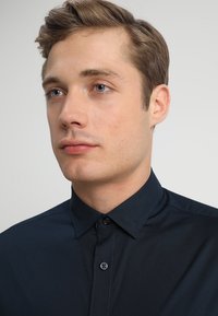 Dunkelblaue button-up Hemd mit spitzem Kragen, minimalistischem Design und kontrastierenden Knöpfen. Glatte Textur, enganliegende Form.