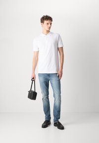Camisa polo blanca, vaqueros azul claros, zapatos negros, sosteniendo un pequeño bolso negro. Corte ajustado, mangas cortas, con un sutil logo en la camisa.