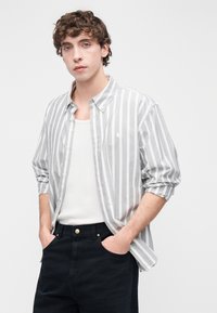 Πουκάμισο με κουμπιά σε ανοιχτό γκρι με λευκές κάθετες ρίγες, συνδυασμένο με λευκό αμάνικο τοπ και σκούρο παντελόνι. Λείο ύφασμα, άνετη γραμμή.