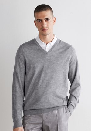 Homme portant un pull à col en V gris clair sur une chemise blanche à col, debout avec une main dans la poche d'un pantalon gris clair assorti.