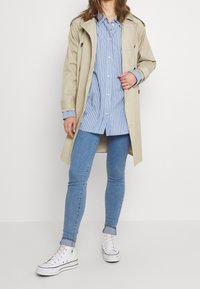 Gabardina beige sobre una camisa de botones a rayas azul y blanca, combinada con jeans ajustados azules y zapatillas blancas.