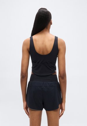 Pikkade juustega naine mustas varrukatutas toppis ja mustades spordipükstes, seistes seljaga vaataja poole tavalise heleda tausta ees.