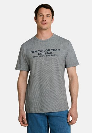 Mann steht nach vorne blickend, trägt ein graues Kurzarm-T-Shirt mit dem Aufdruck "Tom Tailor Team EST 1962" und blaue Jeans vor einem schlichten weißen Hintergrund.