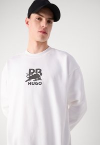 Hvit sweatshirt med ribbestrikket rund hals, med en svart logo av en okse og tekst "PB HUGO" sentrert på brystet. Mykt stoff, avslappet passform.