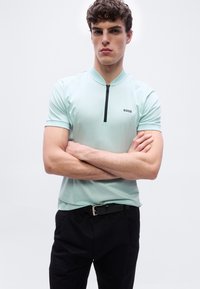 Νεαρός άνδρας με σταυρωμένα χέρια, φορώντας ανοιχτό μπλε polo με φερμουάρ και το λογότυπο "BOSS" καθώς και μαύρο παντελόνι, σε λευκό απλό φόντο.