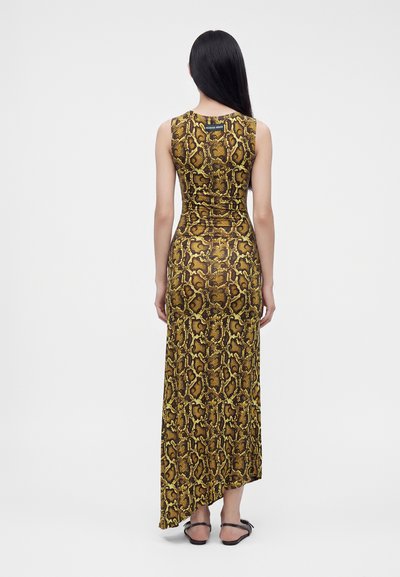 Femme aux longs cheveux noirs portant une robe maxi asymétrique sans manches à imprimé serpent jaune et marron et des sandales noires, de dos.