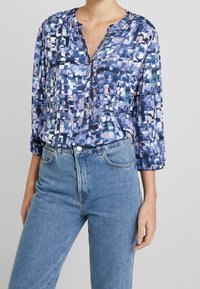 Kolorowa bluzka w wzory w odcieniach niebieskiego i fioletowego, z okrągłym dekoltem, zapięciem na guziki i długimi rękawami. Połączona z dżinsowymi spodniami.