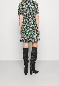 Blumenkleid mit kurzen Ärmeln auf schwarzem Hintergrund, verziert mit weißen Gänseblümchen und grünen Akzenten; kombiniert mit knielangen schwarzen Lederstiefeln.