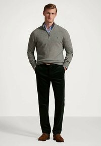 Sivi pulover na zadrgo, prilagojen kroj. Temno zeleni kordurovi hlače, mehka tekstura. Rjavi čevlji, bronast zaponka pasu, stoječi položaj na nevtralnem ozadju.