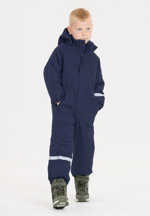 Navyblauwe waterdichte sneeuw overal met een ritssluiting aan de voorkant, capuchon en reflecterende strepen, gecombineerd met groene winterlaarzen met veters.