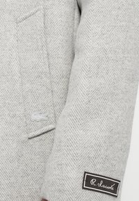 Sivý vzorovaný blejzer z látky herringbone, s malým bielym vyšitým logom, bočnou kapsou a štítkom s nápisom "R. Lacoste" na rukáve.