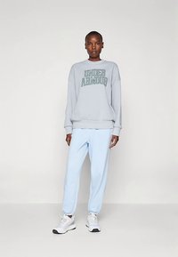 Lichtgrijze sweatshirt met "UNDER ARMOUR" in donkergroen gedrukt, gecombineerd met lichtblauwe joggingbroek en witte sportschoenen. Glad stoffen oppervlak.