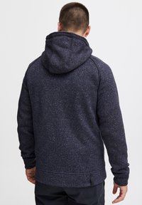 Marineblå hoodie med tekstureret stof, der har en rummelig hætte, lange ærmer og en kant med et lille mærke på siden.