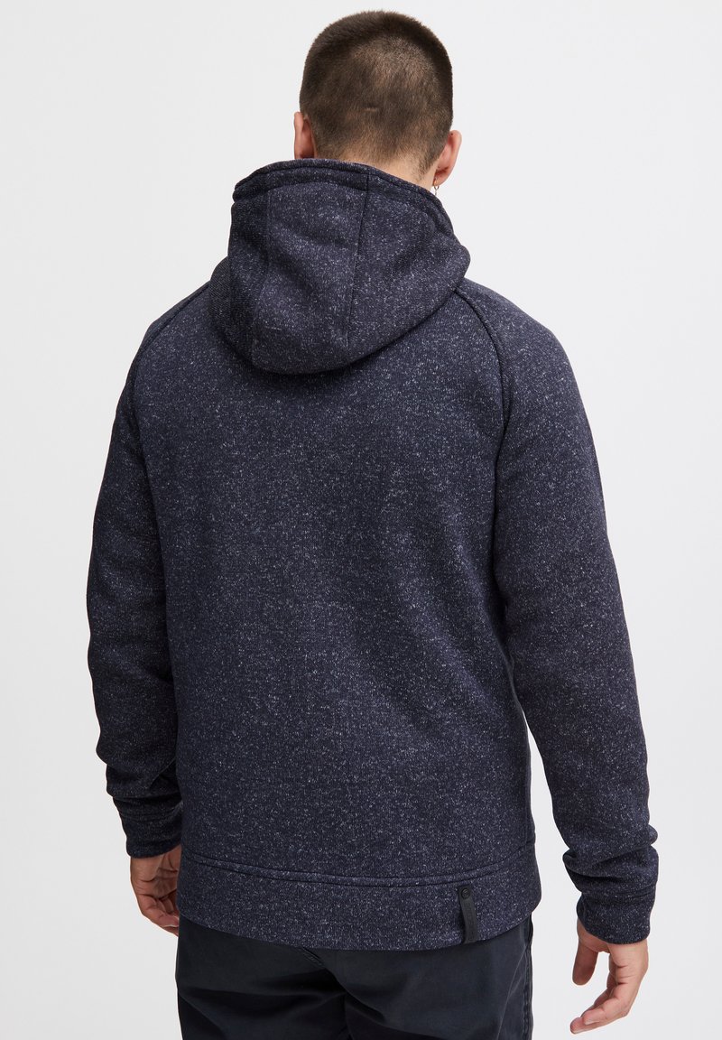 Marineblå hoodie med tekstureret stof, der har en rummelig hætte, lange ærmer og en kant med et lille mærke på siden.