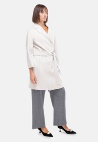 Cappotto avvolgente grigio chiaro con cintura, maniche lunghe e colletto a revers; abbinato a pantaloni grigi larghi e tacchi neri con accenti argentati.