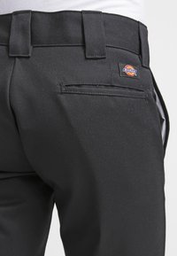Pantalon noir en tissu lisse et résistant, avec une poche arrière et une étiquette du logo Dickies. Le design comprend des passants pour ceinture et une coupe ajustée.
