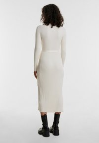 Weißes geripptes Strickkleid mit tailliertem Schnitt und langen Ärmeln. Mit einem gebundenen Taillendetail und Midi-Länge, kombiniert mit schwarzen Knöchelstiefeln.